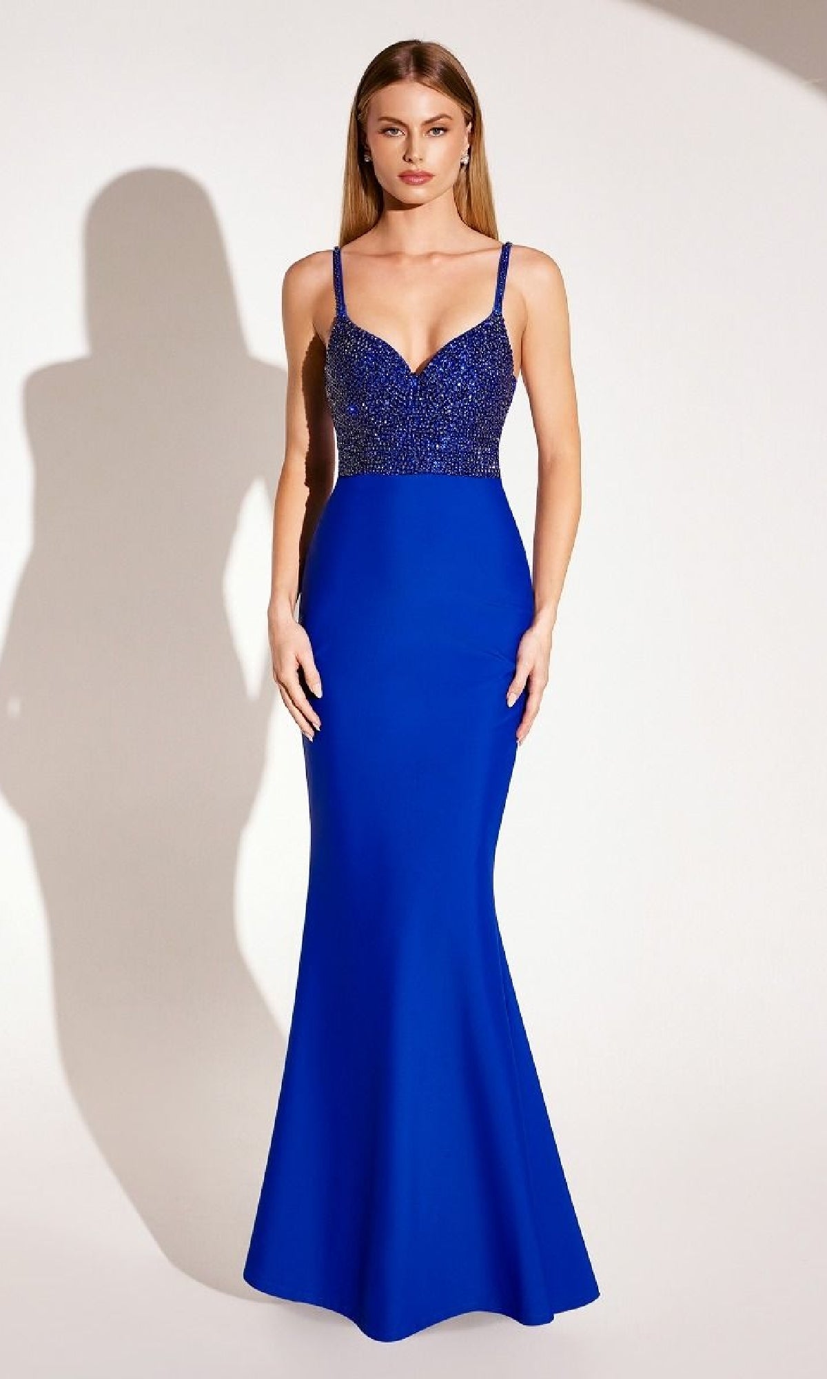 A woman stands against a plain light background wearing the Amoris T9002 Beaded-Bodice Long Prom Dress, a royal blue, floor-length mermaid gown with thin straps.